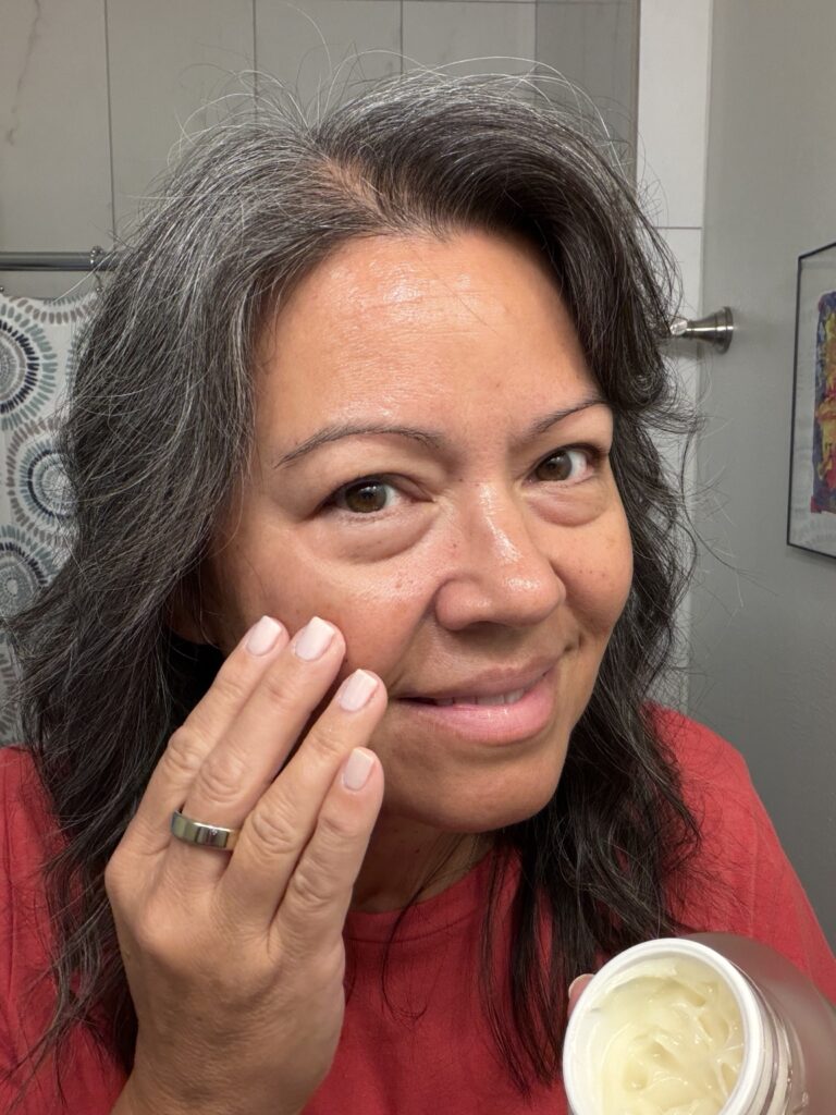 Cindy Ortiz smiling in her bathroom while holding an open jar of face cream during her skincare routine
